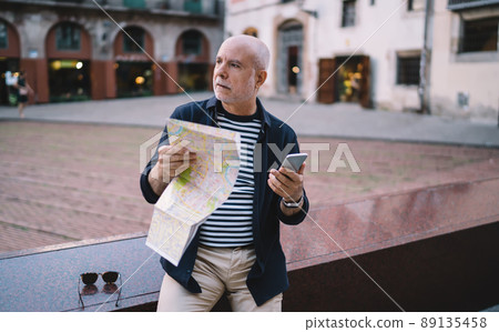 Retired male tourist with travel map and modern cellphone gadget for GPS tracking thinking in city 89135458