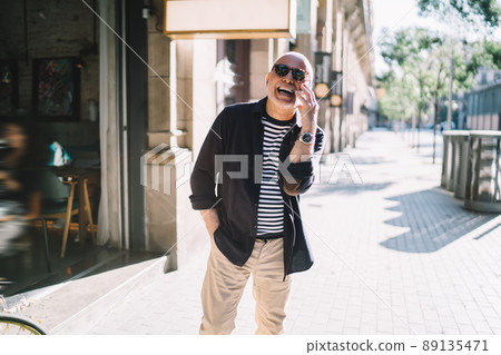 Cheerful elderly man laughing in city while calling Cheerful elderly man laughing in city while calling 89135471