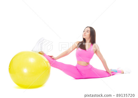 A beautiful, athletic, slim, smiling and cheerful woman in a pink tracksuit demonstrates stretching. Sits on a twine supported by a yellow fitball. Pilates. Lifestyle concept with sports and gym 89135720