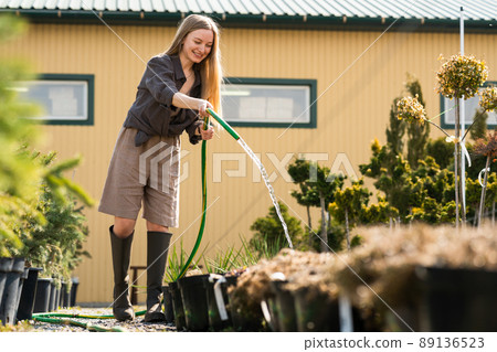Happy female garden center worker watering plant pots with a hose 89136523