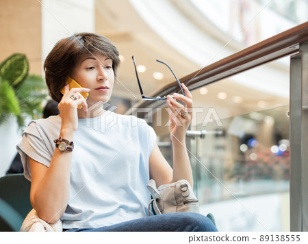 Woman is talking by smartphone and is looking surprisingly on her eyeglasses at shopping mall. Coffee break at store. Woman is talking by smartphone and is looking surprisingly on her eyeglasses at shopping mall. Coffee break at store. 89138555