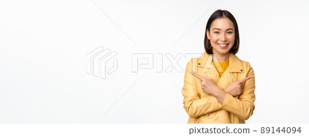 Image of indecisive asian girl, pointing fingers sideways, pointing left and right, choosing variant, deciding, standing over white background 89144094