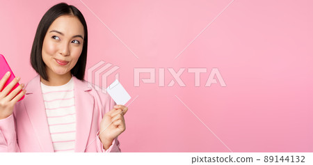 Smiling asian corporate woman, lady in suit thinking, holding smartphone and credit card, plan to buy smth online, shopping with mobile phone, pink background 89144132