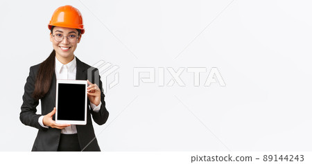 Smiling professional female engineer, saleswoman or estate agent showing location to client, standing in safety helmet and business suit, introduce construction project at tablet screen Smiling professional female engineer, saleswoman or estate agent showing location to client, standing in safety helmet and business suit, introduce construction project at tablet screen 89144243
