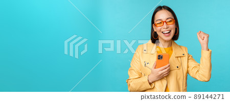 Enthusiastic smiling asian girl in sunglasses, holding mobile phone and dancing, laughing with smartphone, standing over blue background Enthusiastic smiling asian girl in sunglasses, holding mobile phone and dancing, laughing with smartphone, standing over blue background 89144271