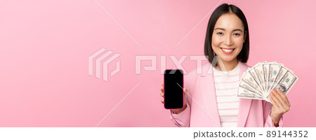 Image of korean successful corporate woman showing money, dollars and smartphone app screen, interface of mobile phone application, concept of investment and finance, pink background 89144352