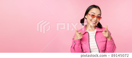 Close up portrait of modern asian girl in sunglasses smiling, pointing fingers at camera, praise you, inviting or complimenting, standing over pink background Close up portrait of modern asian girl in sunglasses smiling, pointing fingers at camera, praise you, inviting or complimenting, standing over pink background 89145072