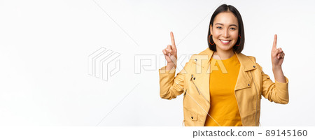 Enthusiastic asian girl pointing fingers up, showing advertisement on top, smiling happy, demonstrating promo offer or banner, standing over white background 89145160