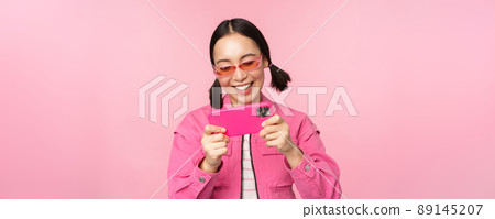 Portrait of happy asian girl playing on smartphone, watching videos on mobile phone app, standing over pink background 89145207