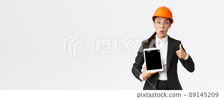 Impressed and excited asian female engineer, industrial technician in business suit and safety helmet showing thumbs-up and digital tablet screen with amazed expression, react to profit diagram 89145209