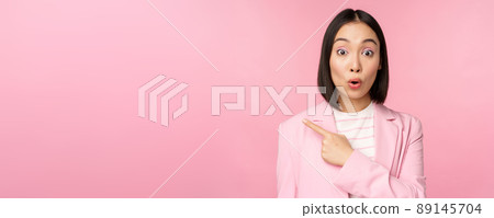 Portrait of businesswoman with surprised face, pointing finger left, showing smth interesting, standing over pink background in office suit 89145704