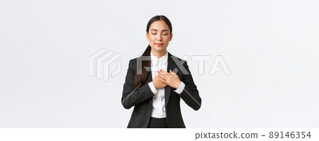 Happy dreamy asian female entrepreneur in suit close eyes and holding hands on heart, remember lovely memory, thinking about something pleasant over white background 89146354