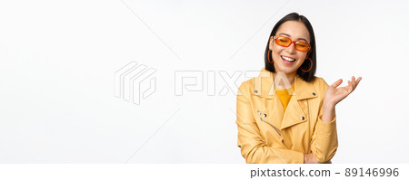 Stylish, modern korean girl in sunglasses and yellow coat, laughing and smiling, happy normal face expression, posing against white studio background Stylish, modern korean girl in sunglasses and yellow coat, laughing and smiling, happy normal face expression, posing against white studio background 89146996