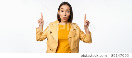 Surprised asian girl pointing fingers up, express interest, showing advertisement ahead, demonstrating promo banner on top, standing against white background 89147003