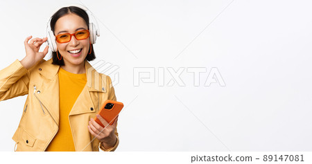 Happy stylish asian woman in sunglasses, listening music in headphones, holding smartphone, singing and dancing, standing over white background 89147081