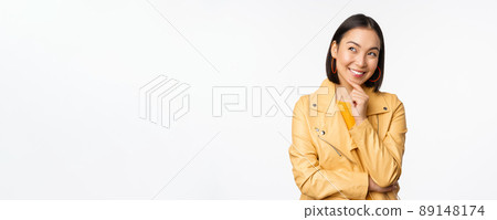 Portrait of stylish korean woman in yellow jacket, smiling thoughtful, thinking and looking up at logo or advertisement, standing over white background 89148174