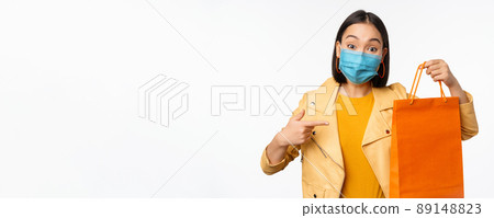 Covid-19 and shop concept. Young asian stylish woman, wearing medical face mask, holding shopping bag, going in malls during pandemic, white background Covid-19 and shop concept. Young asian stylish woman, wearing medical face mask, holding shopping bag, going in malls during pandemic, white background 89148823