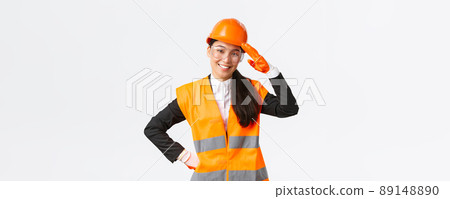 Confident smiling, friendly asian female construction manager, engineer in safety helmet and gloves saluting, greeting architect or chief at enterprise, showing building area to investors 89148890