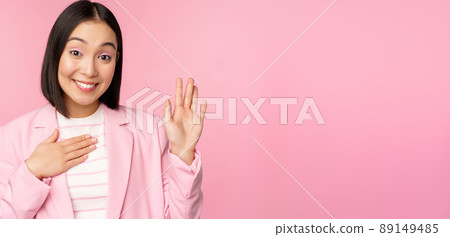 Portrait of cute asian businesswoman raising hand, introduce herself in office, smiling coquettish, standing over pink background in suit Portrait of cute asian businesswoman raising hand, introduce herself in office, smiling coquettish, standing over pink background in suit 89149485