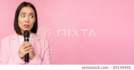 Insecure asian businesswoman giving speech, scared of talking in public, using microphone, standing in suit over pink background Insecure asian businesswoman giving speech, scared of talking in public, using microphone, standing in suit over pink background 89149488