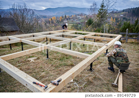 Man building wooden frame house on pile foundation. Male worker drilling hole by electric drill in timber framing of future house on construction site in the mountains. Carpentry concept. 89149729