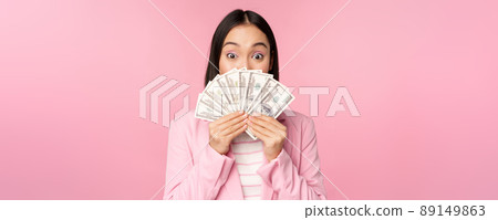 Happy asian lady in suit holding money, dollars with pleased face expression, standing over pink background 89149863
