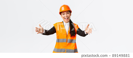 Friendly smiling asian female construction manager, engineer in safety helmet and reflective jacket, extending hands for greeting, welcome business partners, standing white background 89149881