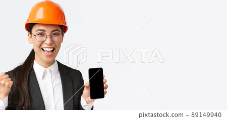 Successful winning asian female engineer, architect in safety helmet and suit, showing smartphone screen, fist pump and shouting yes from rejoice, triumphing over achievement, white background Successful winning asian female engineer, architect in safety helmet and suit, showing smartphone screen, fist pump and shouting yes from rejoice, triumphing over achievement, white background 89149940