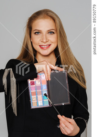 Beautiful middle-aged woman makeup artist holds shadows in her hands and looks at the camera smiling. Blond hair and a black jacket on a light background. Beautiful middle-aged woman makeup artist holds shadows in her hands and looks at the camera smiling. Blond hair and a black jacket on a light background. 89150779