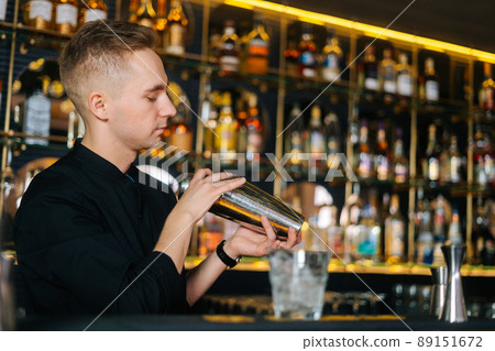 Side view of bartender mixing ingredients of alcoholic cocktail by shaking shaker standing behind bar counter, on blurred background of shelves with alcoholic drinks, selective focus. 89151672