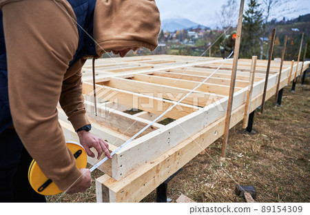 Man worker building wooden frame house on pile foundation. Carpenter using tape measure for measuring wooden planks. Carpentry concept. 89154309