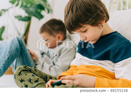 Cheerful two Childs boys playing online game, watching video on cellphone. Smiling kids using funny mobile apps, enjoying free leisure time at home on couch. Brothers play together at phone 89155236