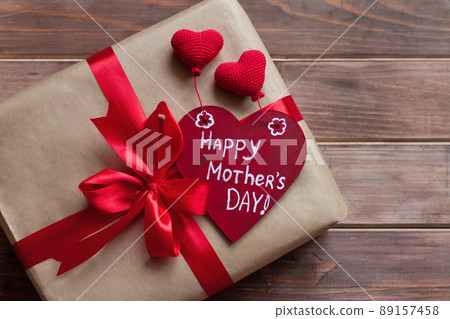 gift in brown craft paper with red ribbon with bow and wooden red heart with white inscription happy mother's day, knitted hearts on old brown table gift in brown craft paper with red ribbon with bow and wooden red heart with white inscription happy mother's day, knitted hearts on old brown table 89157458