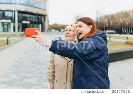 Happy Mother and adult positive daughter are doing Selfie on city street, Urban lifestyle concept. Travelers. Outdoor shot of well-dressed female friends. 89157626