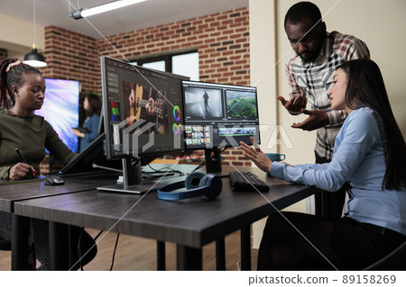 Footage editing specialist using multi monitor workstation to improve frames quality. Professional videographer sitting at desk while coworker helping her with film visual improvement. Footage editing specialist using multi monitor workstation to improve frames quality. Professional videographer sitting at desk while coworker helping her with film visual improvement. 89158269