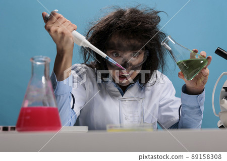 Foolish silly female lab worker with pipette and glass jar mixing toxic chemical compounds to create new formula. Crazy mad scientist mixing liquid substances while grinning dreadful. 89158308