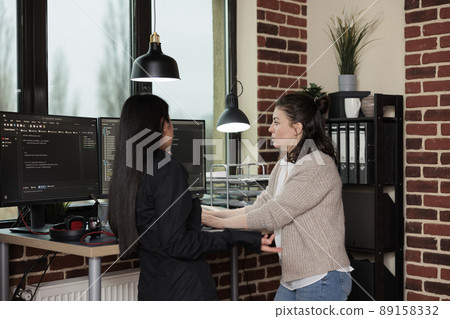Company programmers standing near desk with multiple monitors on top of it. Network developers updating firewall system through information processing using multiple layers of security. Company programmers standing near desk with multiple monitors on top of it. Network developers updating firewall system through information processing using multiple layers of security. 89158332