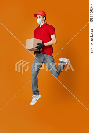Delivery man in face mask holding cardboard box and jumping Delivery man in face mask holding cardboard box and jumping 89160920
