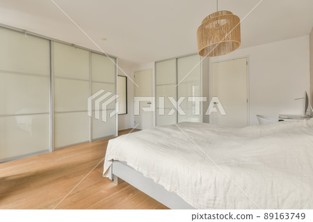A spacious bedroom with a large white bed, a huge wardrobe and a workplace with a computer in a modern house A spacious bedroom with a large white bed, a huge wardrobe and a workplace with a computer in a modern house 89163749