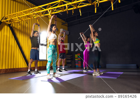 Group of young people training at gym, doing stretching and yoga exercises indoors. Warming up exercises before training session Group of young people training at gym, doing stretching and yoga exercises indoors. Warming up exercises before training session 89165426
