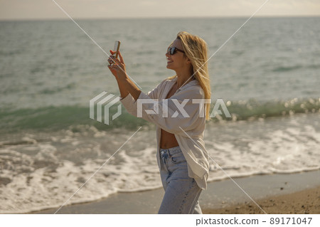 Side view of female tourist taking pictures of the sea using smartphone, standing on the coast 89171047