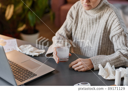 High angle shot of unrecognizable woman having flu wearing sweater sitting at desk at home holding glass with mixture of water and effervescent tablet High angle shot of unrecognizable woman having flu wearing sweater sitting at desk at home holding glass with mixture of water and effervescent tablet 89171129