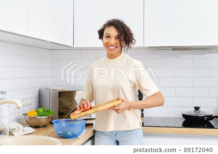 Young african american housewife drops chopped tomato into the bowl in the kitchen 89172040