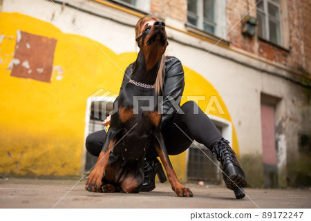 A girl in leather, with a Doberman, in an old yard. 89172247