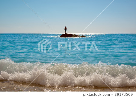 Waves breaking on the sandy beach. The man stands on a small rocky island by the sea and getting ready to jump into the water. 89173509
