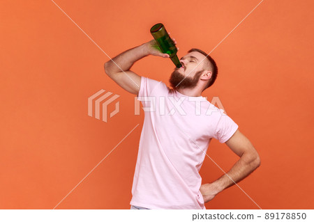 Portrait of bearded man standing with beer bottle, being drunk, standing with closed eyes and drinking alcohol, wearing pink T-shirt. Indoor studio shot isolated on orange background. 89178850