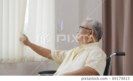 Old asian man sits in a wheelchair and looks out the window 89179815
