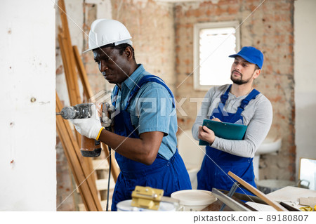 Builder drills a wall with a hammer drill, and his colleague makes notes Builder drills a wall with a hammer drill, and his colleague makes notes 89180807