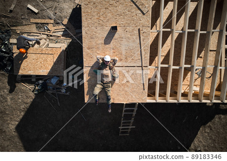 Portrait of male worker building wooden frame house. Top view man laying on rooftop of building on construction site in white helmet. Drone view Portrait of male worker building wooden frame house. Top view man laying on rooftop of building on construction site in white helmet. Drone view 89183346