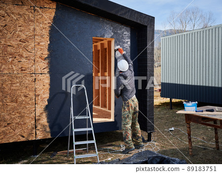 Male painter using paint roller, doing exterior paint work in a black color. Man worker building wooden frame house. Carpentry and construction concept. Male painter using paint roller, doing exterior paint work in a black color. Man worker building wooden frame house. Carpentry and construction concept. 89183751